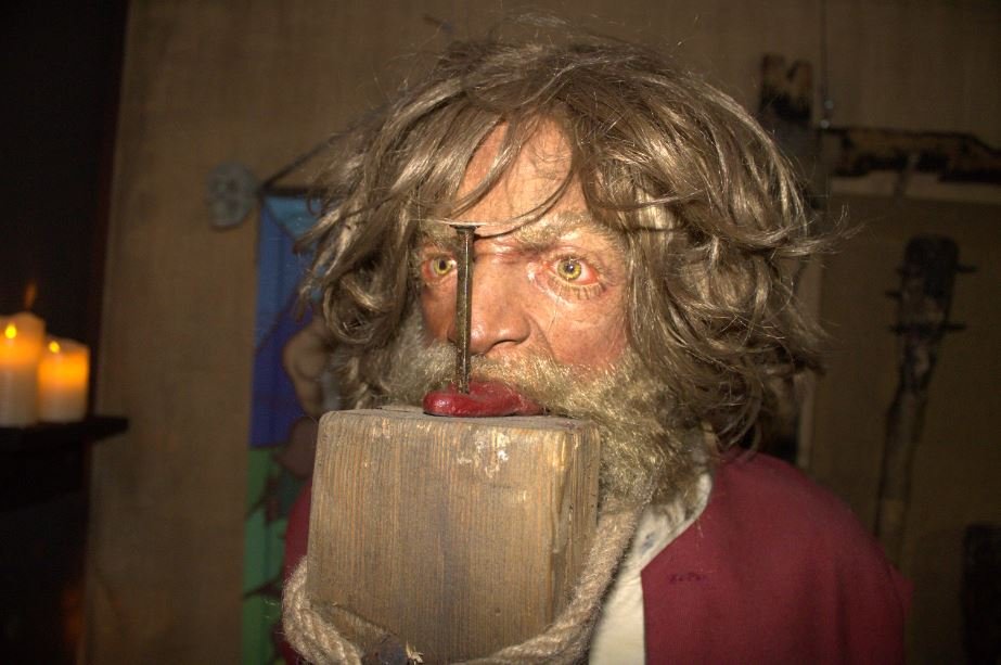 A person with bushy hair and beard, holding a wooden block in front of their face with a nail driven through it. Candles are visible on the left side of the background.