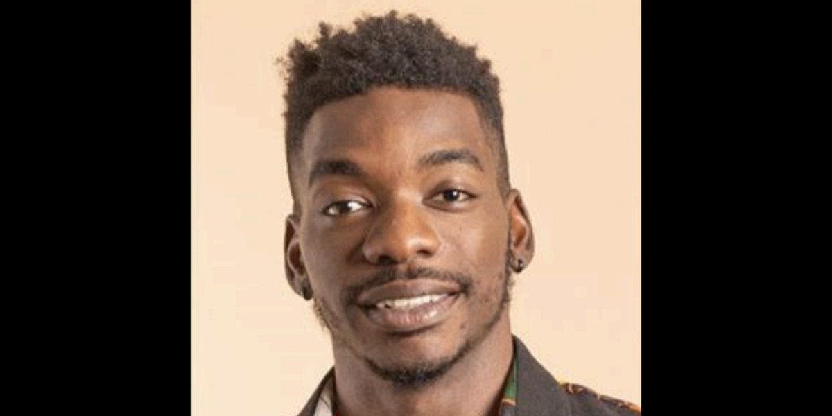 Portrait of a young man with short, curly hair, earrings, a trimmed beard, and a slight smile, wearing a patterned shirt.