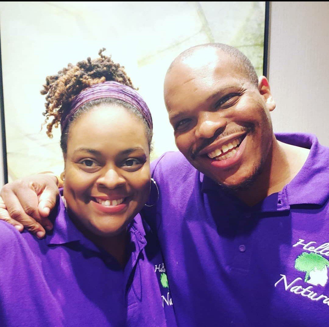 Two people smiling and wearing matching purple shirts, one woman and one man, with the woman having her arm around the man's shoulder.