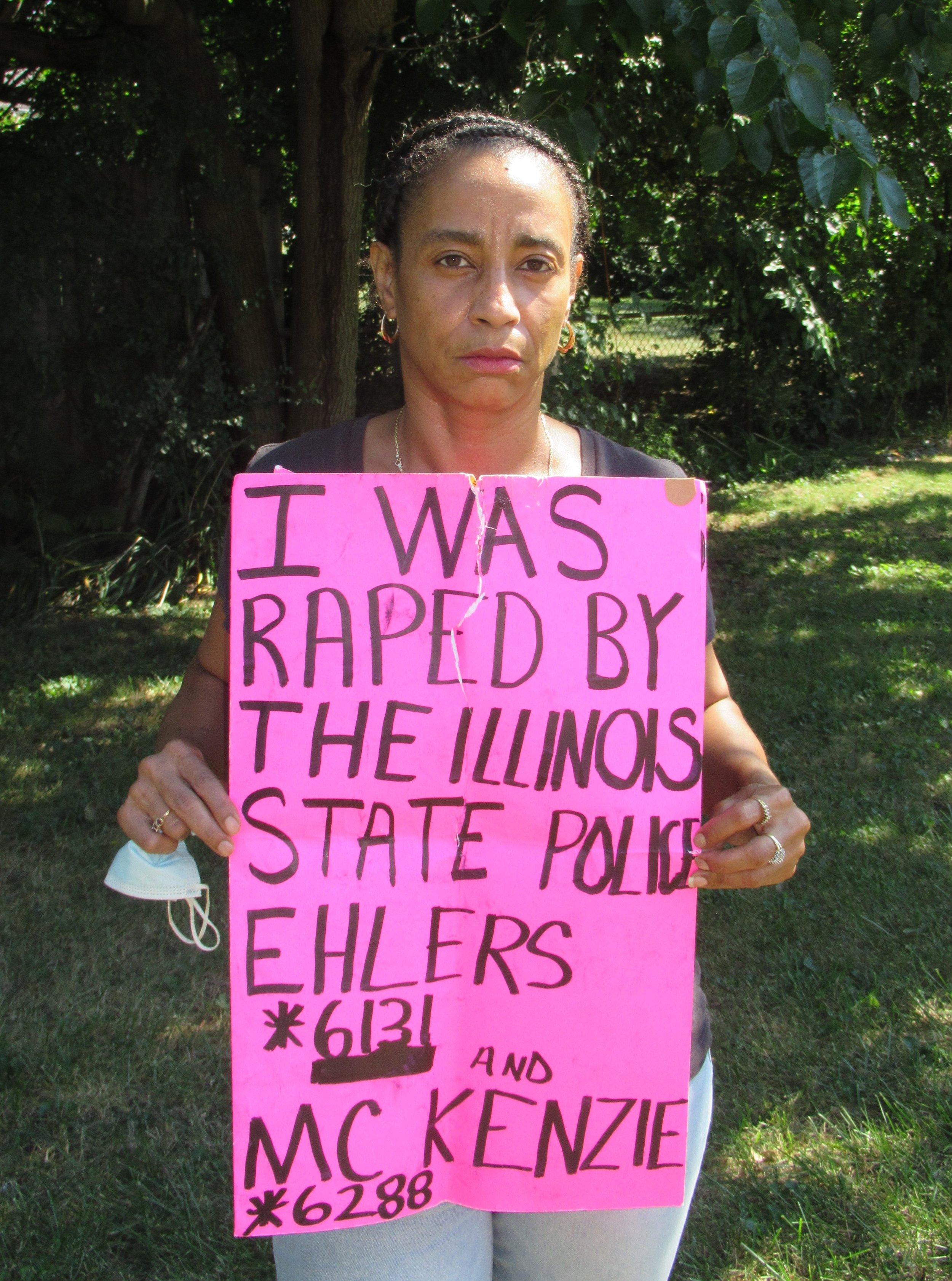 A woman is standing outdoors holding a pink protest sign that reads "I WAS RAPED BY THE ILLINOIS STATE POLICE EH LERS 6131 AND MCKENZIE 6288." She has a serious expression, braided hair, and is wearing a black shirt, earrings, and a necklace. She has a face mask hanging from her hand.