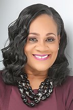 Portrait of a woman with shoulder-length black curly hair, wearing a maroon blazer and a statement black necklace, smiling against a plain light background.