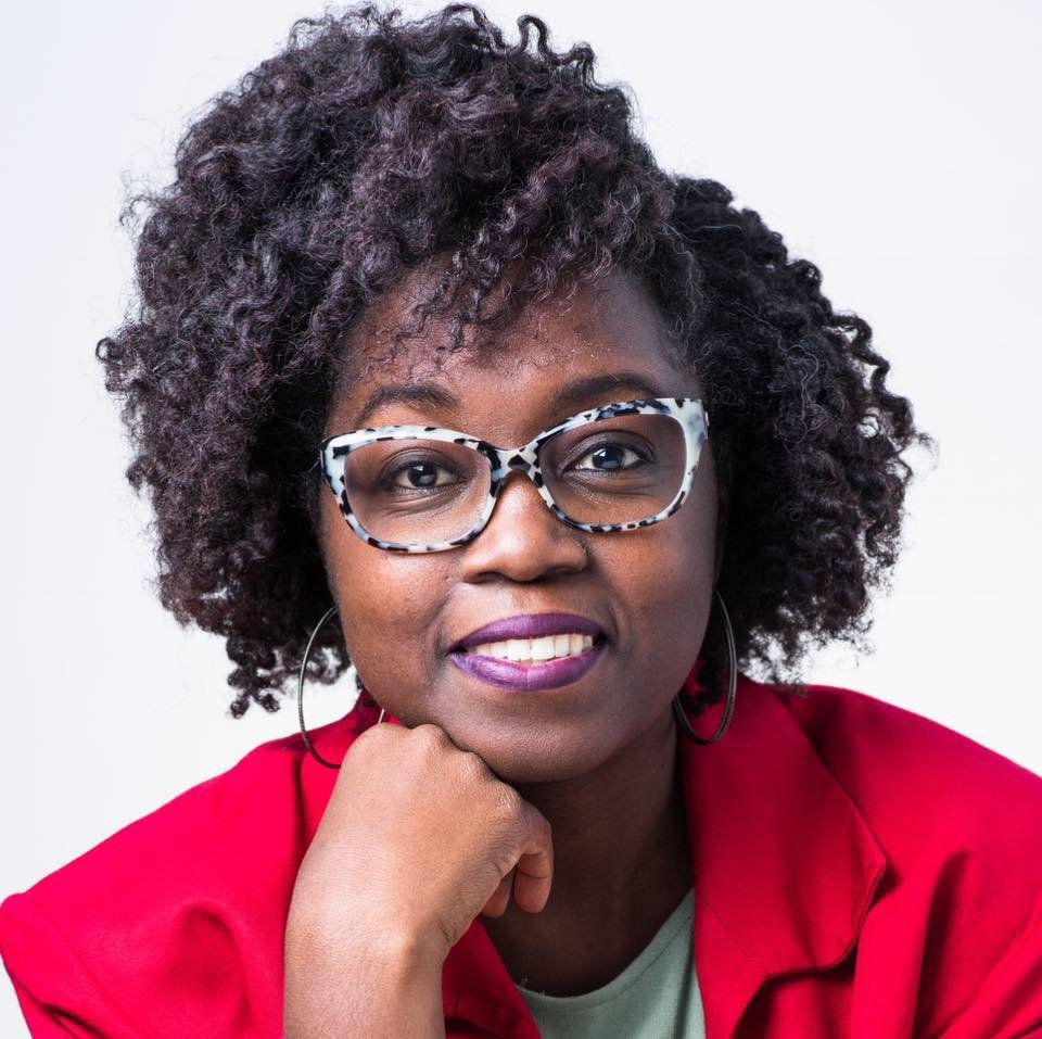 A woman with curly hair wearing glasses and a red jacket, resting her chin on her hand and smiling.