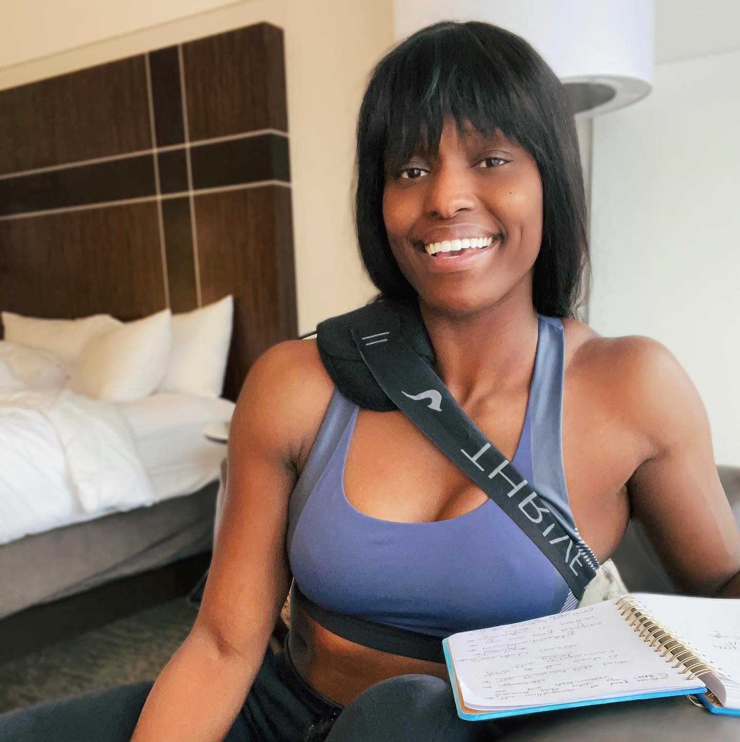 Smiling woman with short black hair, wearing a blue sports bra and a black strap across her shoulder, sitting in a hotel room with a bed in the background, holding a notebook with handwritten notes.