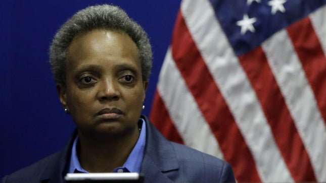 A woman with short gray hair and a serious expression, dressed in a dark blazer and blue shirt, standing in front of an American flag.