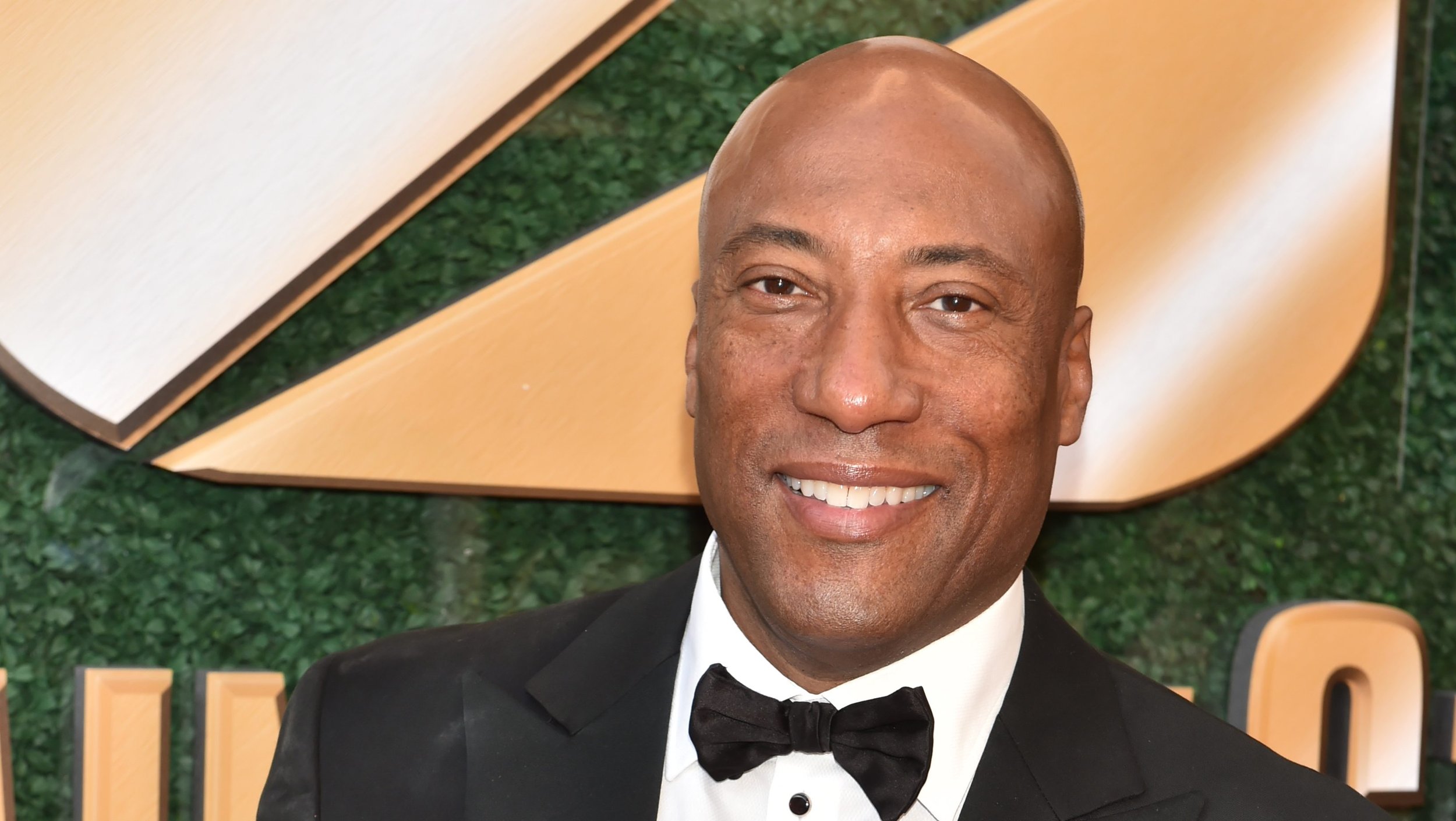 A smiling man wearing a black tuxedo with a black bow tie, standing in front of a green textured background with a large wooden geometric sculpture.