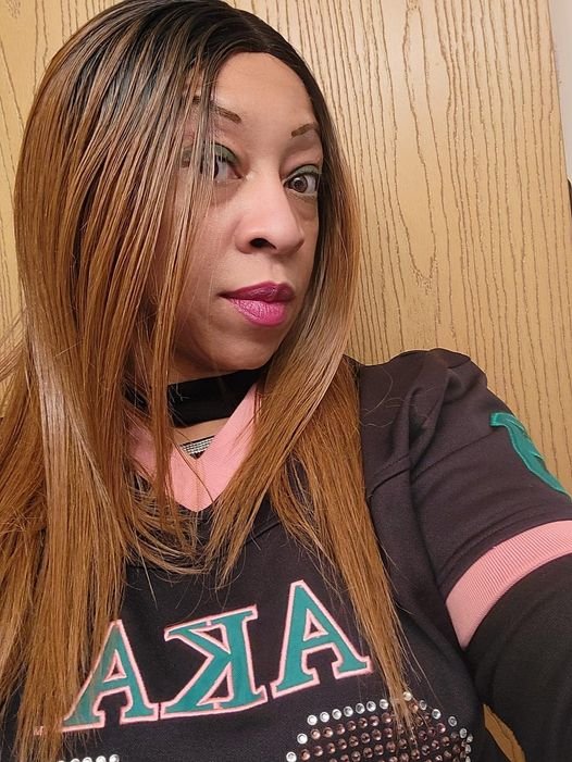 A woman with long, straight, light brown hair and makeup taking a selfie, standing against a wooden wall, wearing a black sports jersey with pink accents and Greek letters.