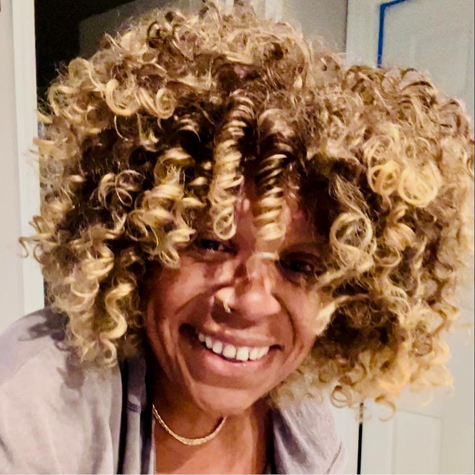 Close-up of a woman with light skin and a wide smile, showing braces, wearing a large, voluminous, blonde curly hairstyle.