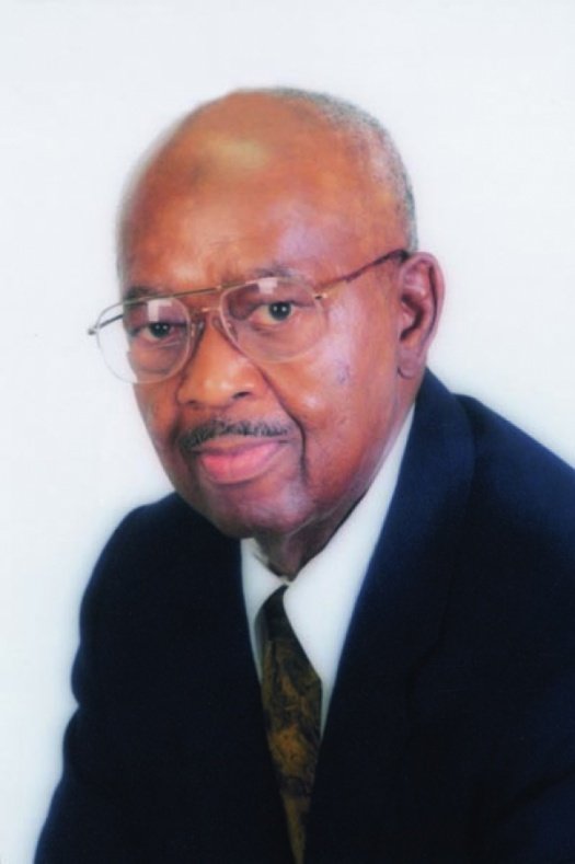 An elderly man with glasses, wearing a navy blazer, white shirt, and tie, sitting against a white background.