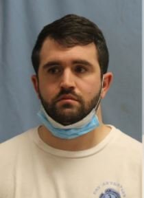 Man with short dark hair and beard wearing a light blue face mask pulled down around his neck, looking to the side, in front of a blue background.