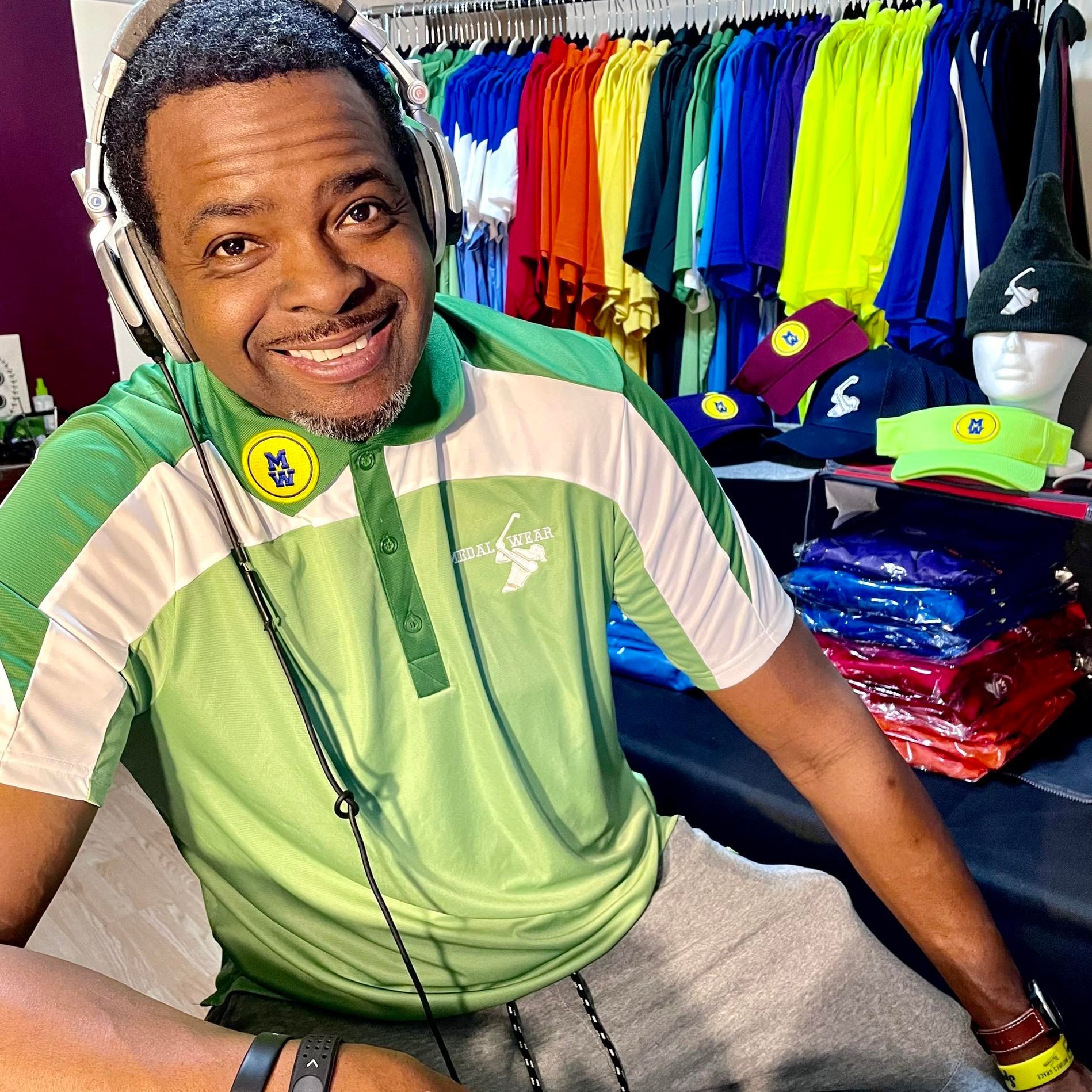 A smiling man wearing a green and white polo shirt with a headset, sitting at a table with colorful sports apparel and accessories, including hats and headbands, in the background.