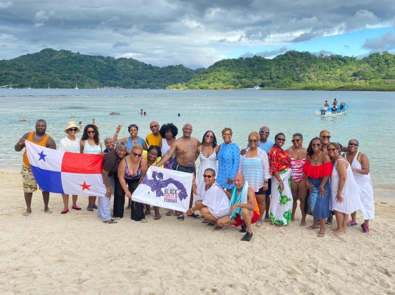 Group of people posing on a beach with water and green mountains in the background. Some are holding flags, with one flag displaying a red, white, and blue design and another with a map of Panama and the text "Black Expats in Panama." The group is dressed in casual, summer clothing, and some are wearing sunglasses and hats. A boat is visible on the water in the background.