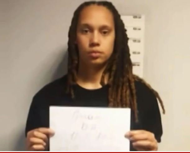 Young woman with long dreadlocks holding a white sign, standing in front of a height chart on a gray wall.