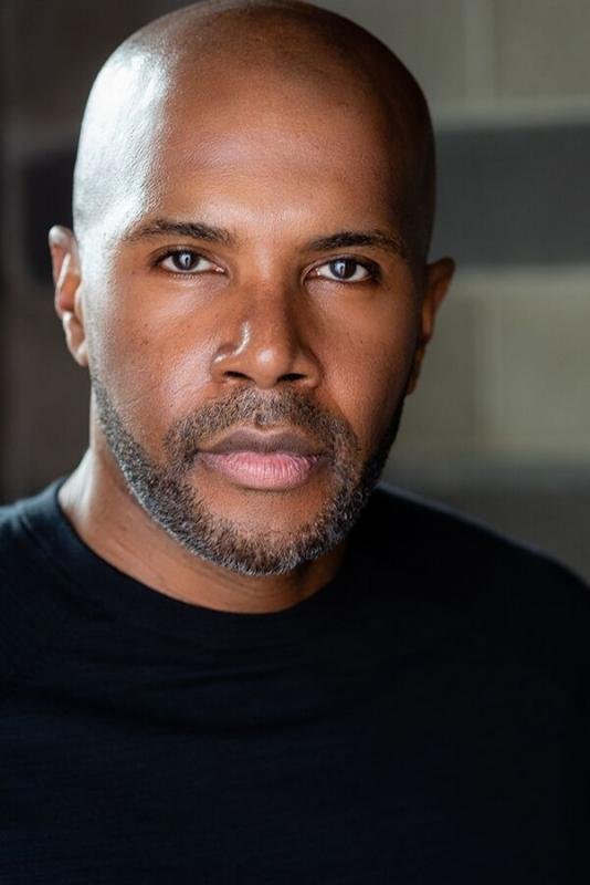 Close-up portrait of a bald African American man with a short beard, wearing a black shirt, looking directly at the camera with a serious expression.