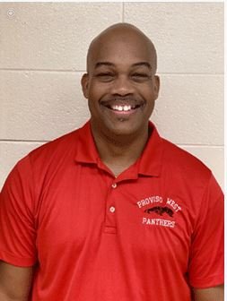 Smiling man in a red polo shirt with university emblem, standing against a white wall.