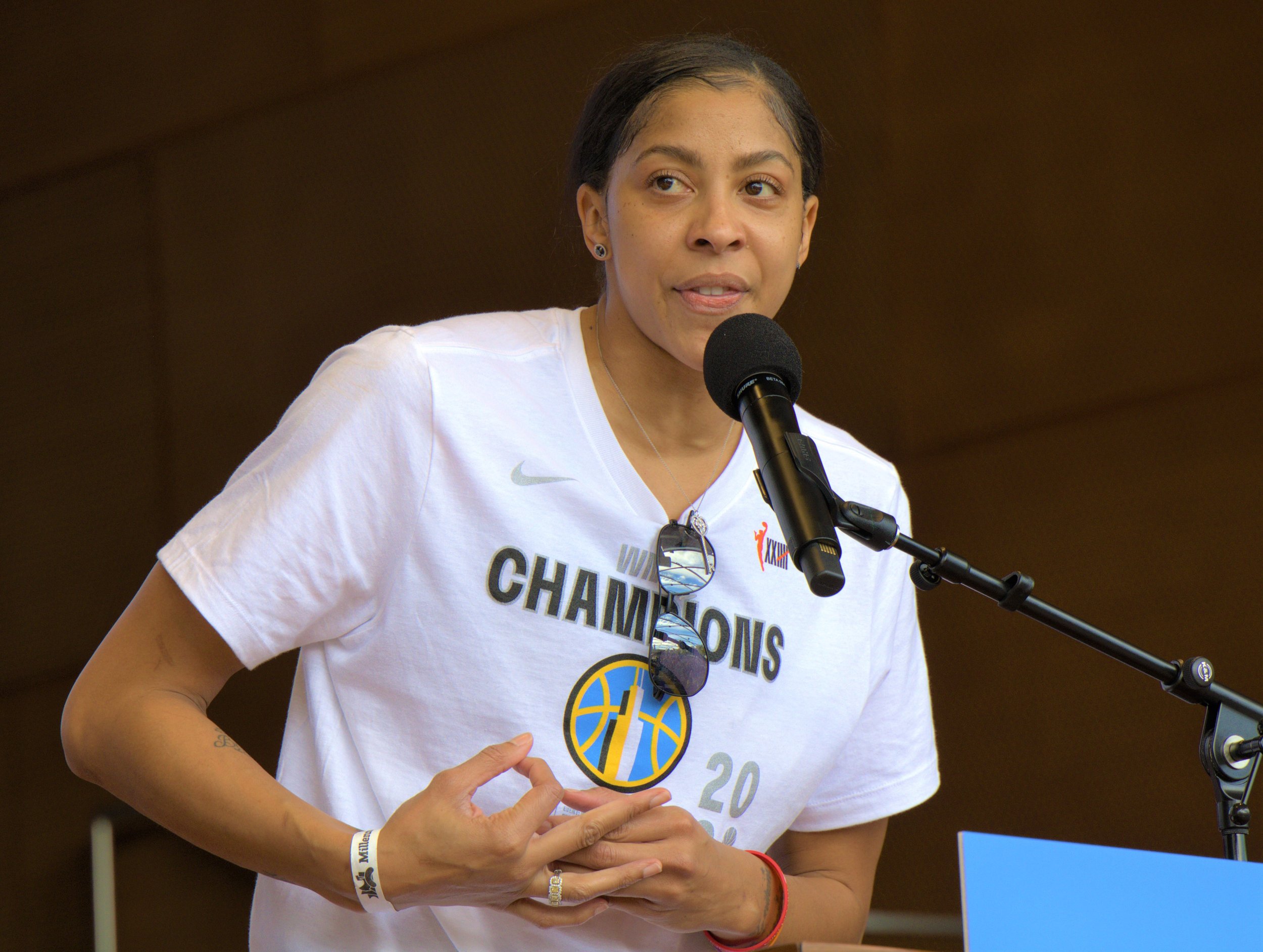 A woman speaking into a microphone, wearing a white shirt with "Champions" written on it, sunglasses hanging from her shirt, and various jewelry, standing indoors.