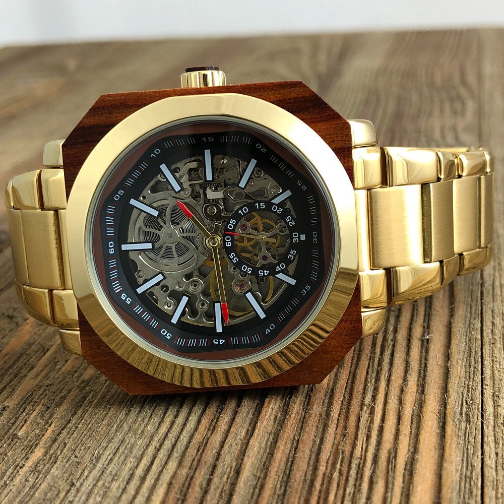 A luxury wristwatch with a gold metal band, a wooden bezel, and a skeleton dial showcasing inner mechanical components, placed on a wooden surface.