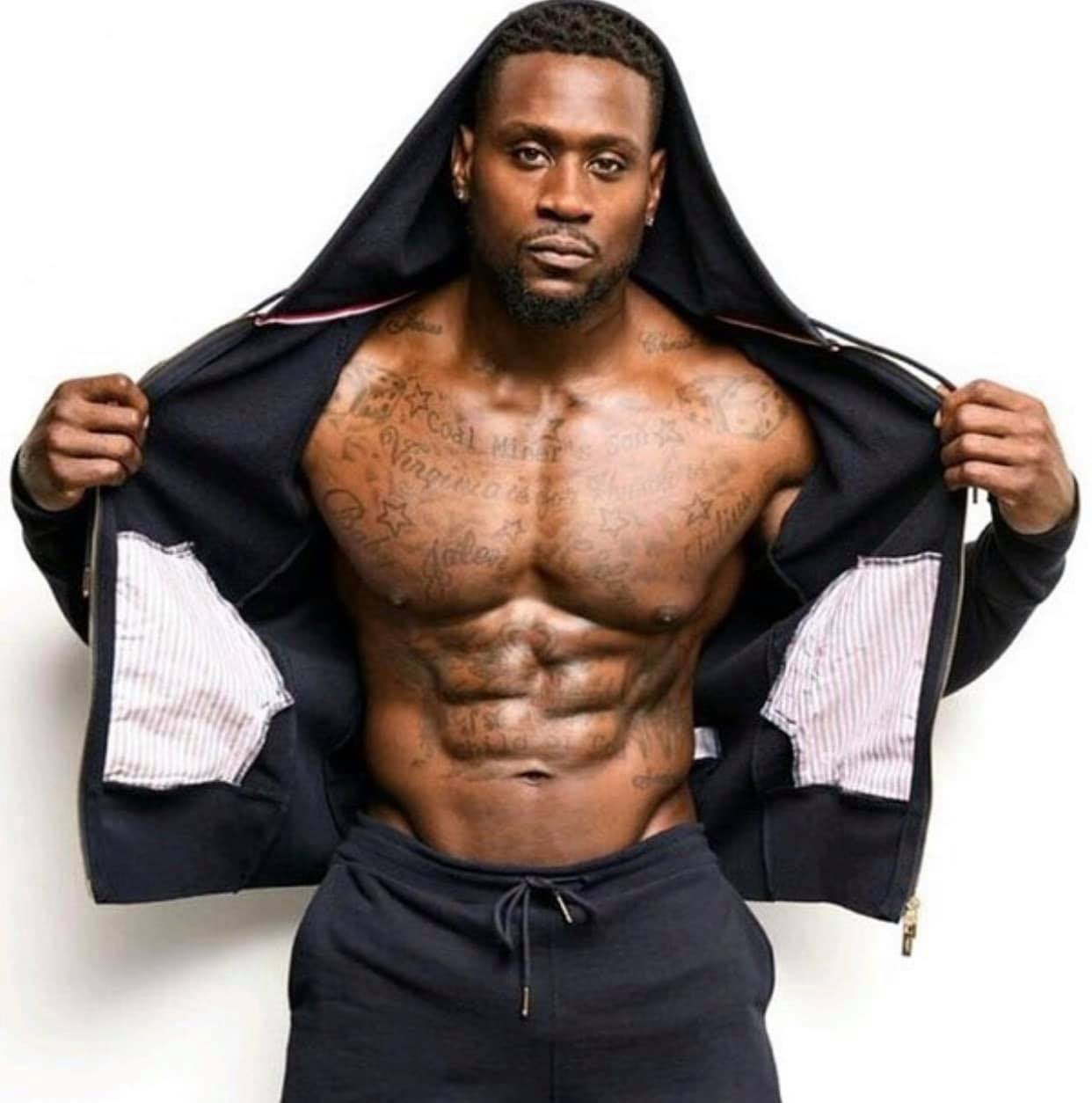 A shirtless man with tattoos pulling open a black jacket to reveal a muscular abdomen against a plain white background.