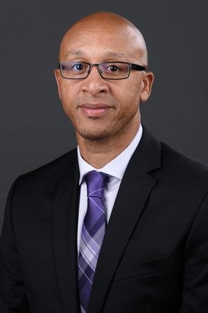 Professional headshot of a man with glasses in a black suit, purple tie, and white shirt against a gray background.