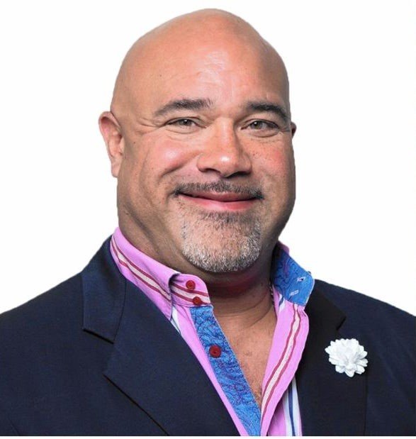 A smiling man with a bald head and goatee, wearing a dark blazer over a pink and purple shirt with a flower pin on the jacket lapel.