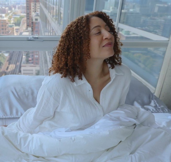 A woman with curly hair sitting in a bed, wearing a white shirt, with her eyes closed and a relaxed expression, in a high-rise apartment with large windows showing a cityscape.