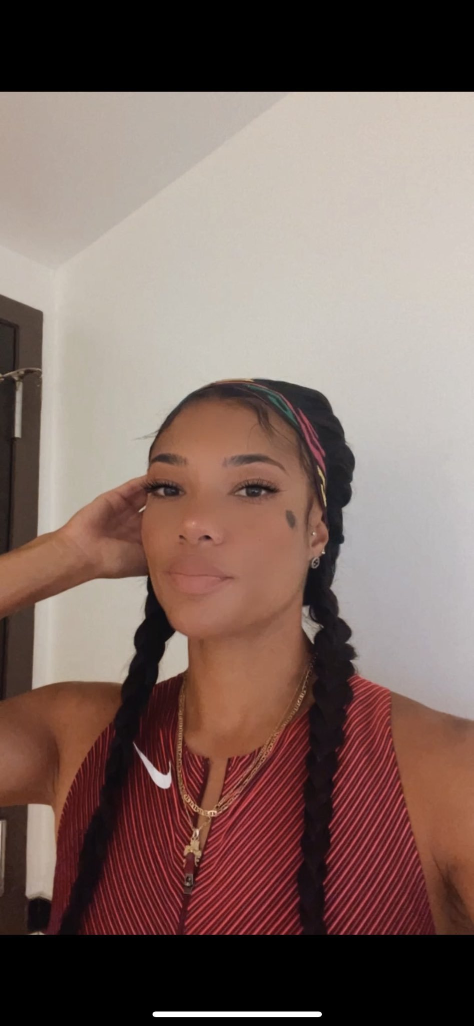 A woman with long black braided hair wearing a red athletic tank top, gold necklaces, and earrings, posing indoors with her hand resting on her head.
