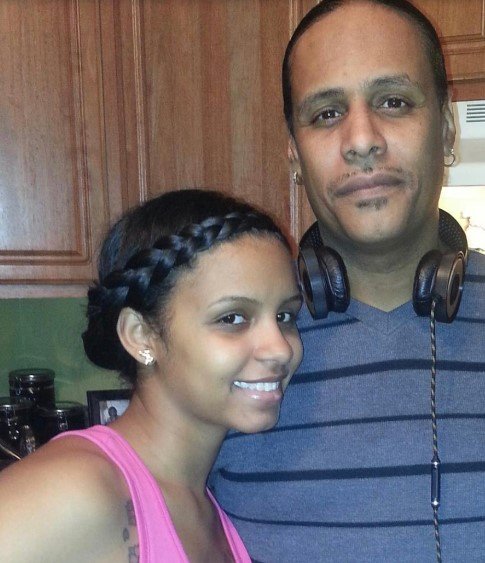 A young woman and an older man posing inside a kitchen, with the woman smiling and the man wearing headphones around his neck.