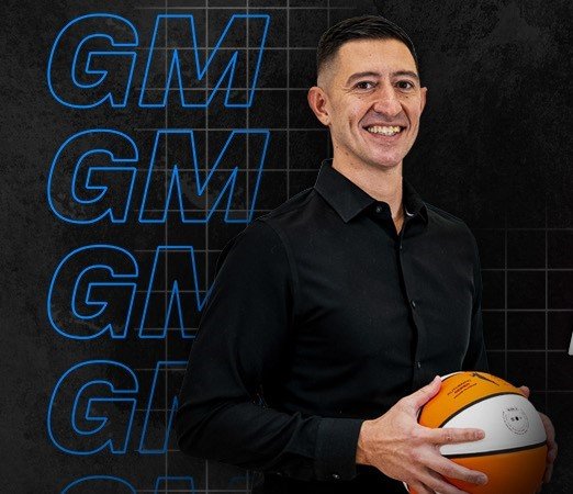 A man in a black shirt holding a basketball, standing in front of a black wall with blue neon 'GM' logos.