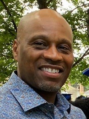 A smiling man in a patterned button-up shirt outdoors with trees in the background.