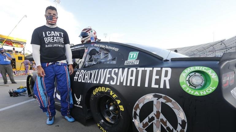 A race car driver in a suit with a helmet, standing next to a black race car with #BlackLivesMatter and other decals, at a racetrack with people and equipment in the background.
