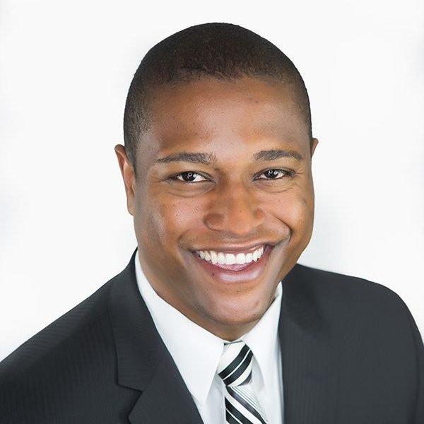 A young man with a short haircut and dark skin, smiling in a black suit, white shirt, and striped tie against a white background.