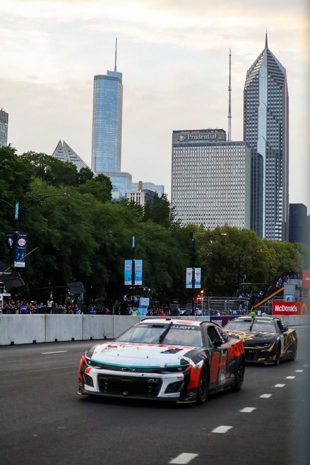 Two race cars speeding on a city street with a downtown skyline in the background, including tall buildings and trees lining the street.