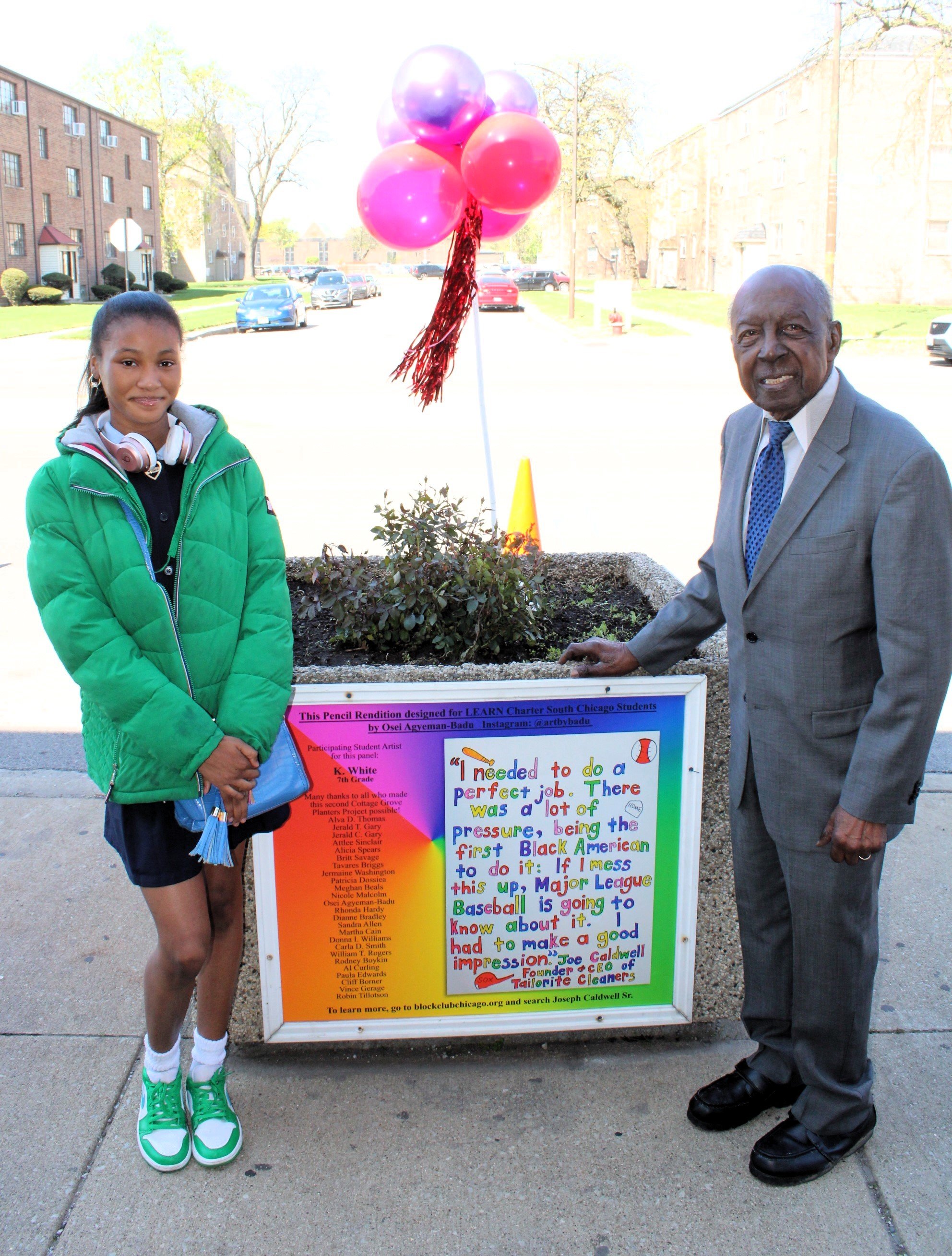 A young girl and an older man standing beside a colorful sign on a sidewalk in a residential neighborhood. The girl is wearing a green jacket, black skirt, and green sneakers, while the man is dressed in a gray suit. Behind them, there is a small tree, pink and purple balloons tied to the tree, and a bright, sunny day with houses and parked cars in the background.