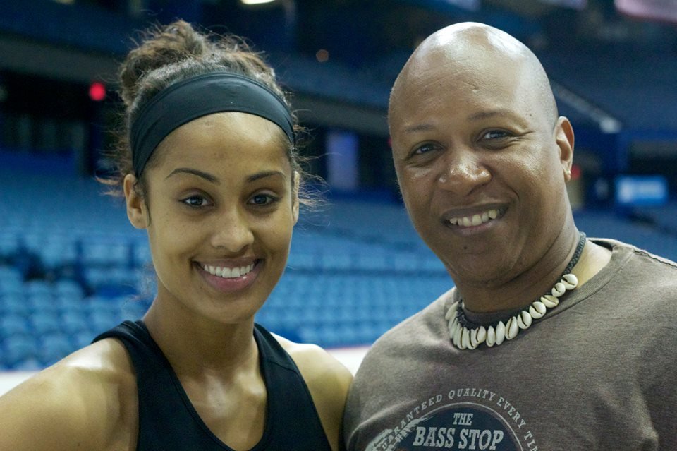 A smiling young woman with a headband and a man with a shaved head wearing a shell necklace, standing together in an indoor sports arena.