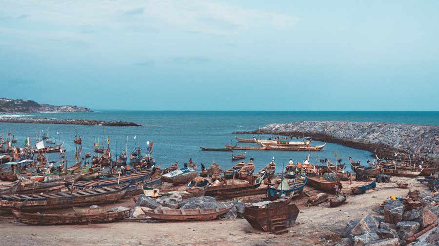 A rocky harbor filled with numerous small boats on the beach, with a breakwater extending into the water and calm sea beyond, under a partly cloudy sky.