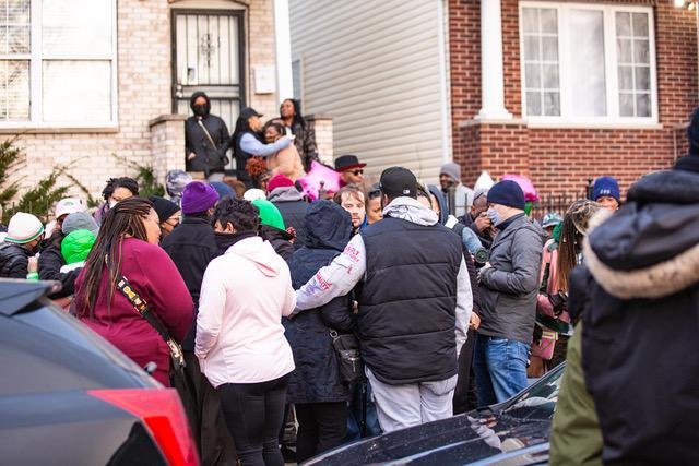 A crowd of people gathers outside a building, some wearing hats and jackets. Two individuals are on a porch, with one holding a baby, seemingly addressing or speaking to the crowd.