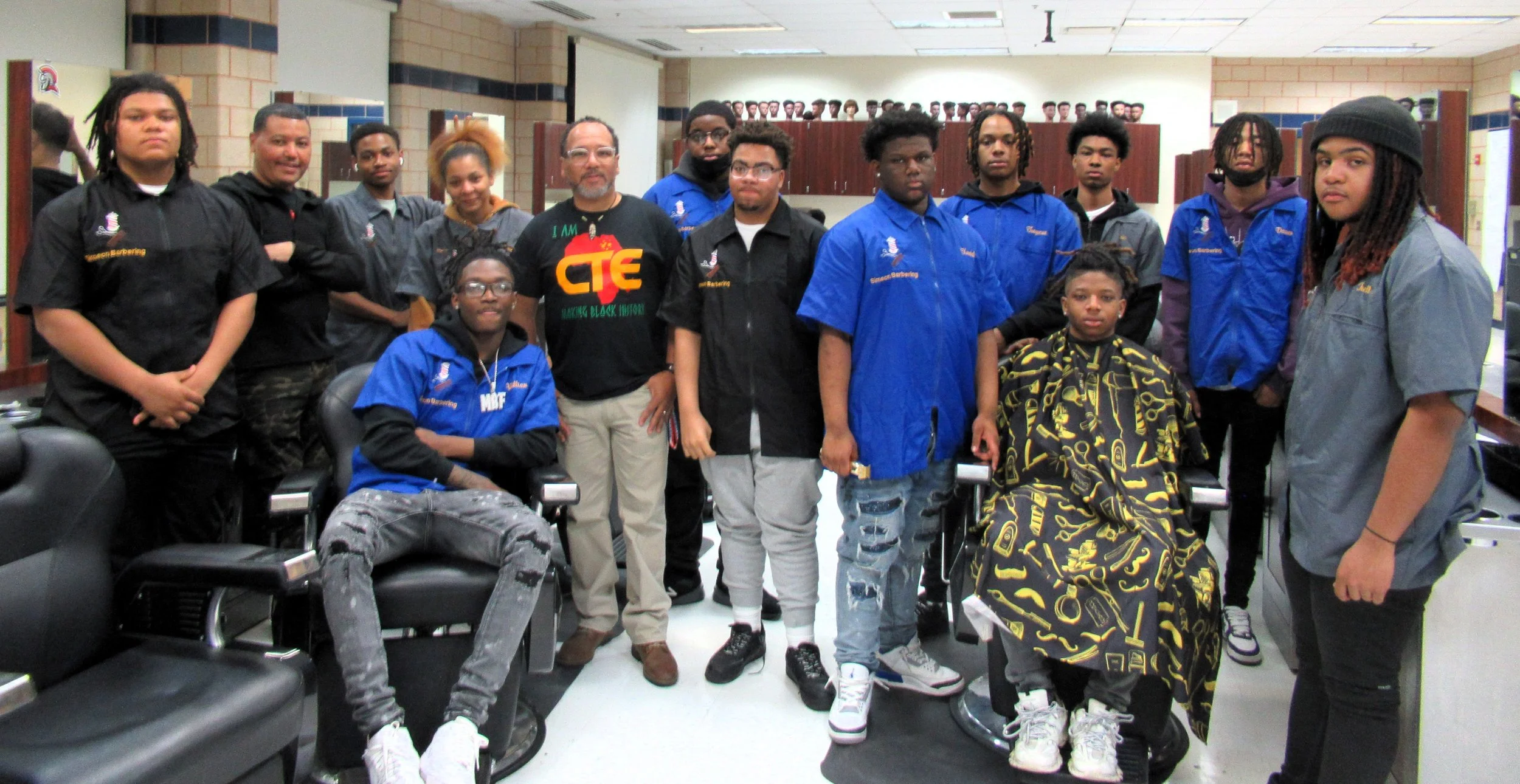 Group of young people and adults in a barbershop setting, with two individuals seated in barber chairs and the others standing behind them, inside a classroom or community space.