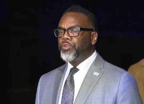 A man with glasses and a beard wearing a light gray suit, white shirt, and tie, speaking at an event.