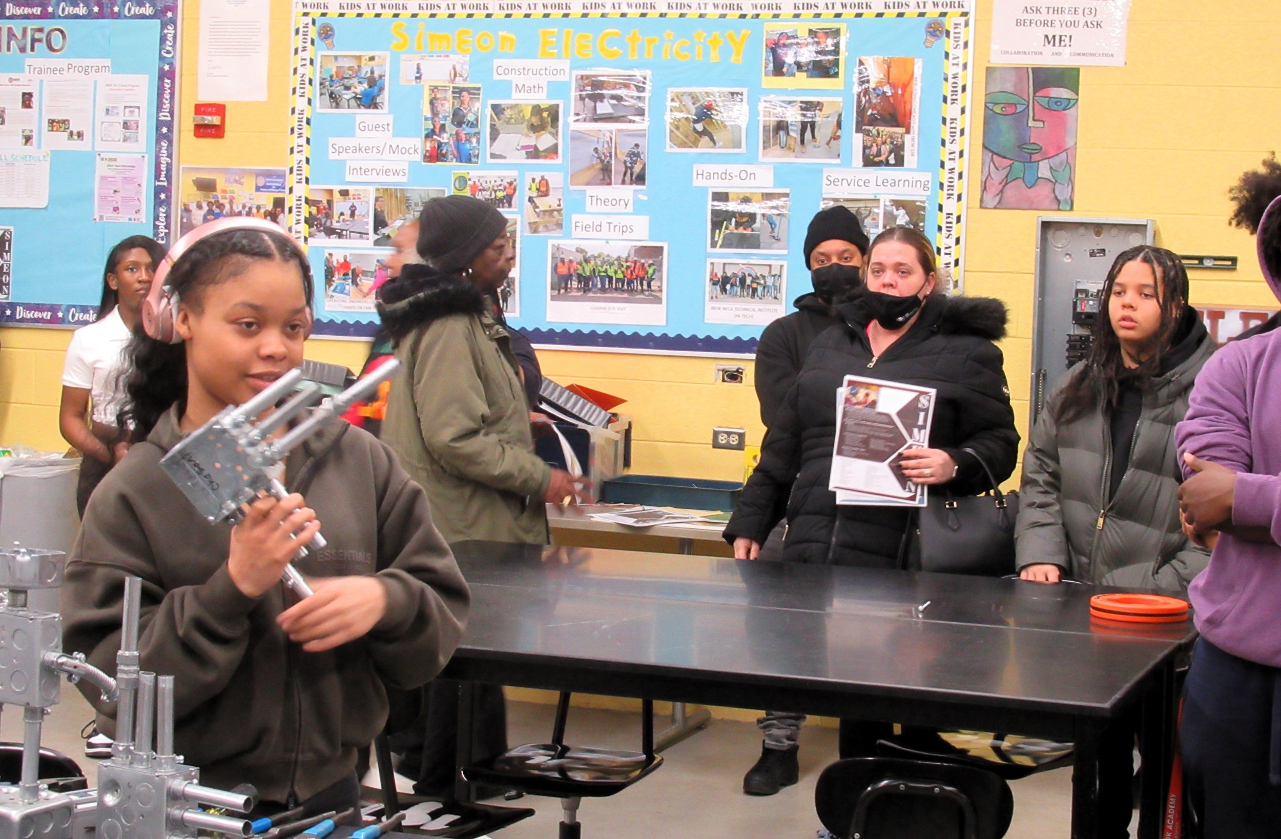 Young girl wearing pink headphones demonstrating a mechanical device in a classroom or workshop setting, with adults observing nearby, children and colorful bulletin boards in the background.
