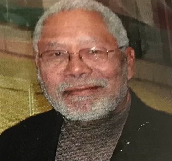 A close-up photo of an older man with gray hair, beard, glasses, and a warm smile, wearing a dark blazer over a brown turtleneck.