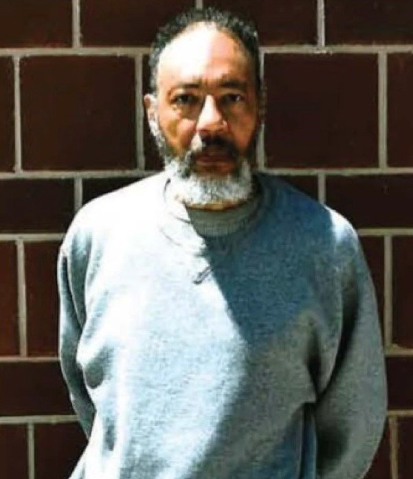 A man with gray hair and beard wearing a gray sweatshirt standing in front of a brick wall.