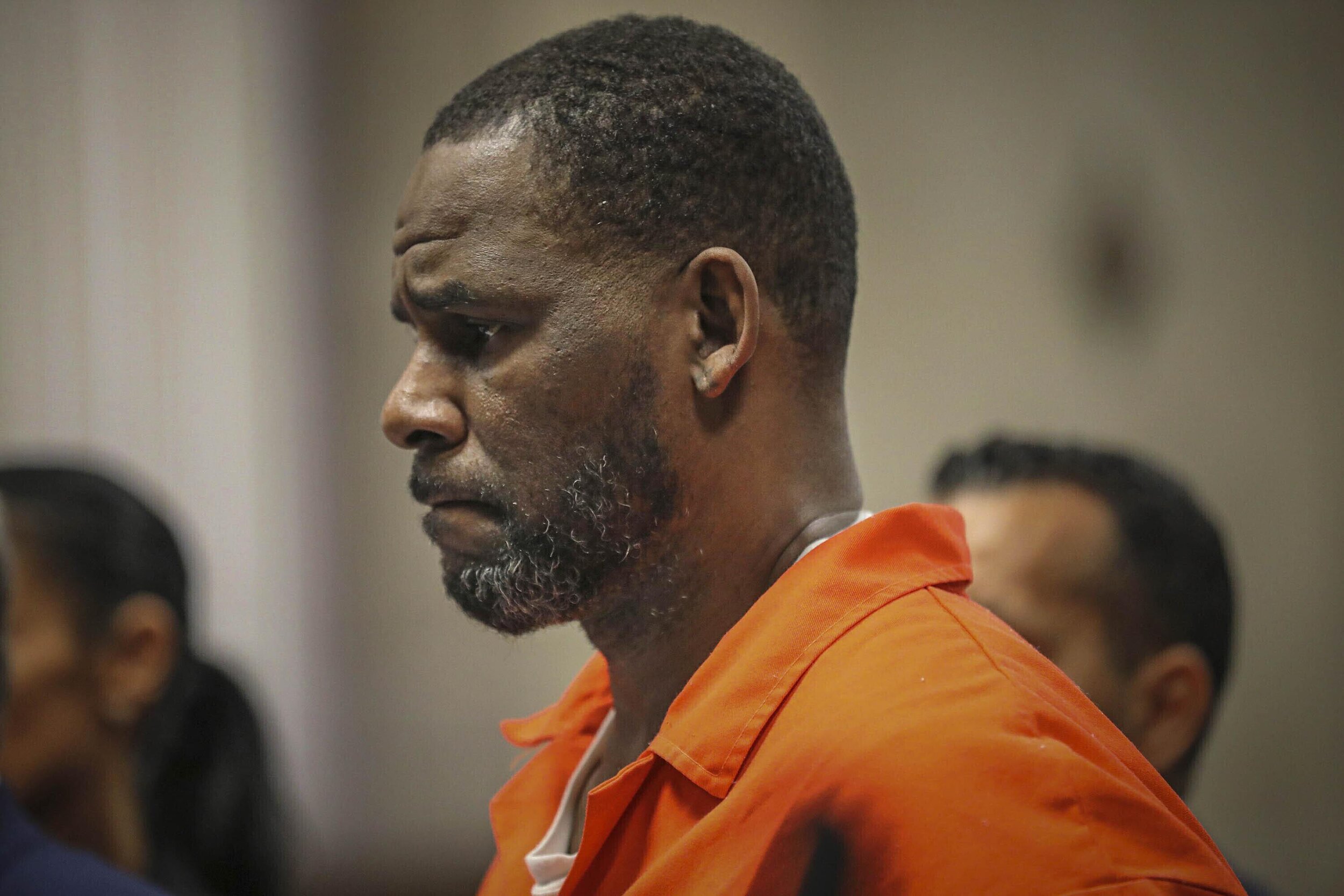 A man wearing an orange prison jumpsuit standing in a room with other people, looking down with a serious expression.