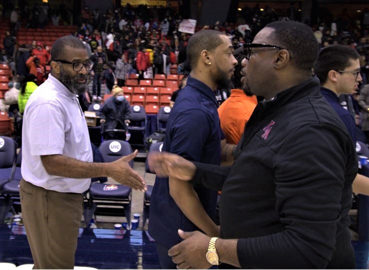 Several men are standing near a basketball court, engaging in conversation and handshakes. The background shows empty seats and other people, suggesting a sports event or game.