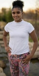 Young girl standing outdoors with hands on hips, wearing a white T-shirt and patterned pants, smiling at the camera.