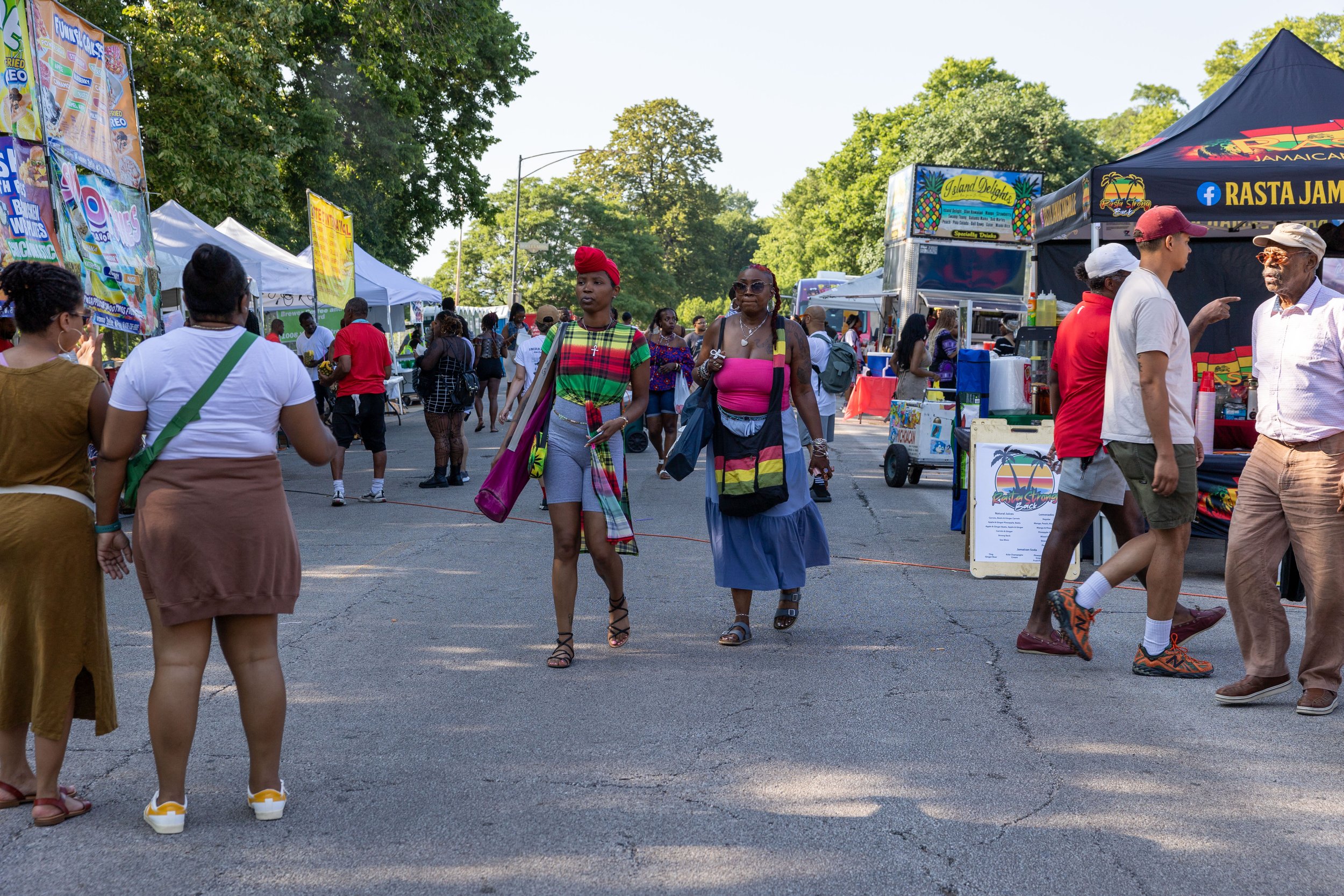 People walking at an outdoor street fair with vendor tents in the background, some trees, and a clear sky.