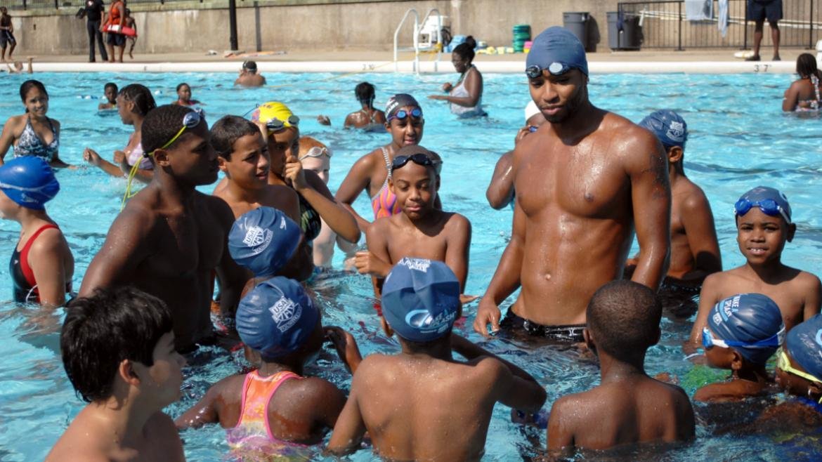 Swim coach giving instructions to a group of children in an outdoor swimming pool.