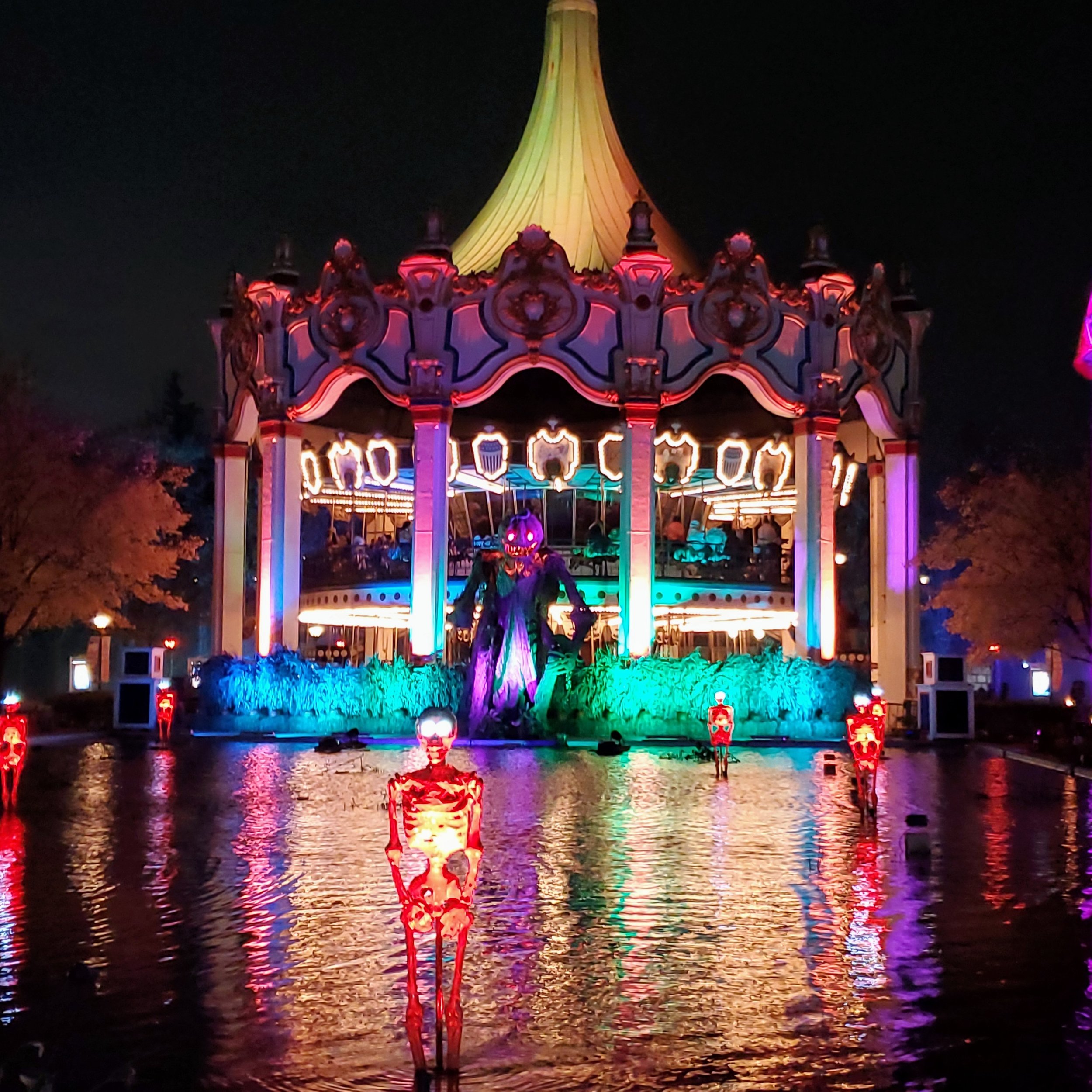 Colorful amusement park carousel lit up at night, featuring skeleton decorations and a pumpkin-headed character in front, reflected in the water.