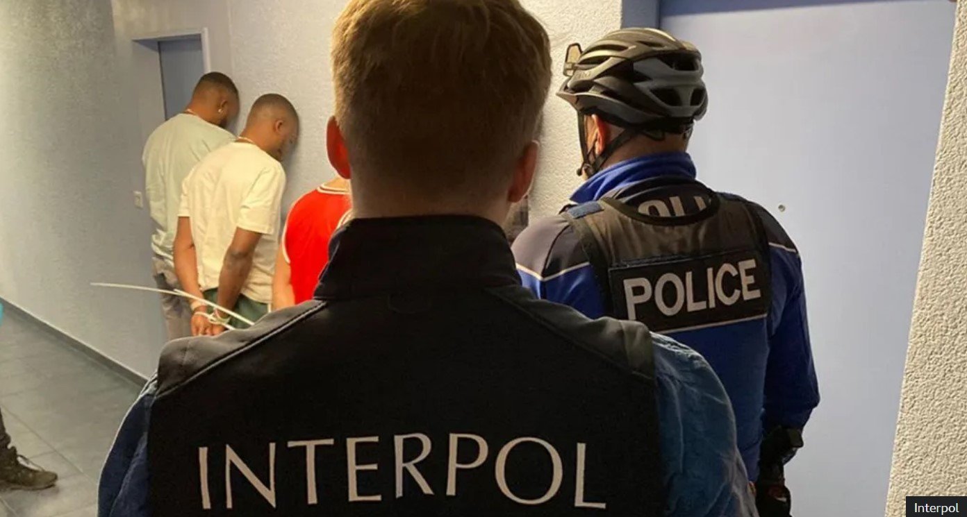 Two police officers, one wearing an Interpol uniform and the other in a police vest with a helmet, stand behind four individuals with their hands against the wall during a security check or arrest in a hallway.