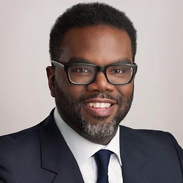 A man with glasses in a dark suit and tie, smiling for a professional photo against a gray background.