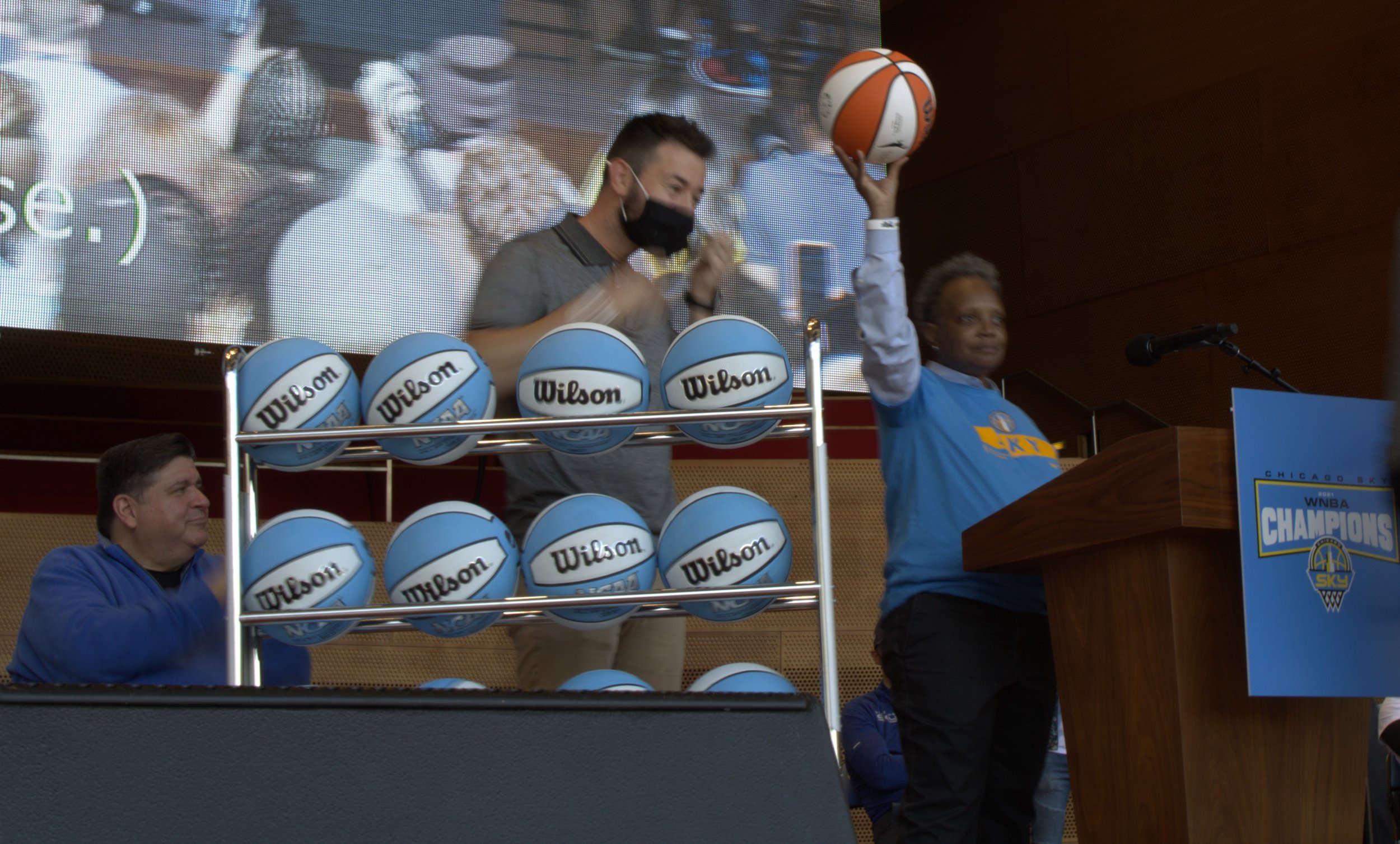 A woman in a blue jersey raises a basketball during a celebration ceremony at the Chicago Sky WNBA Championship event, with a man in a gray shirt and black mask standing behind her and another man in a blue jacket sitting to the left. There are multiple Wilson basketballs on a display rack.