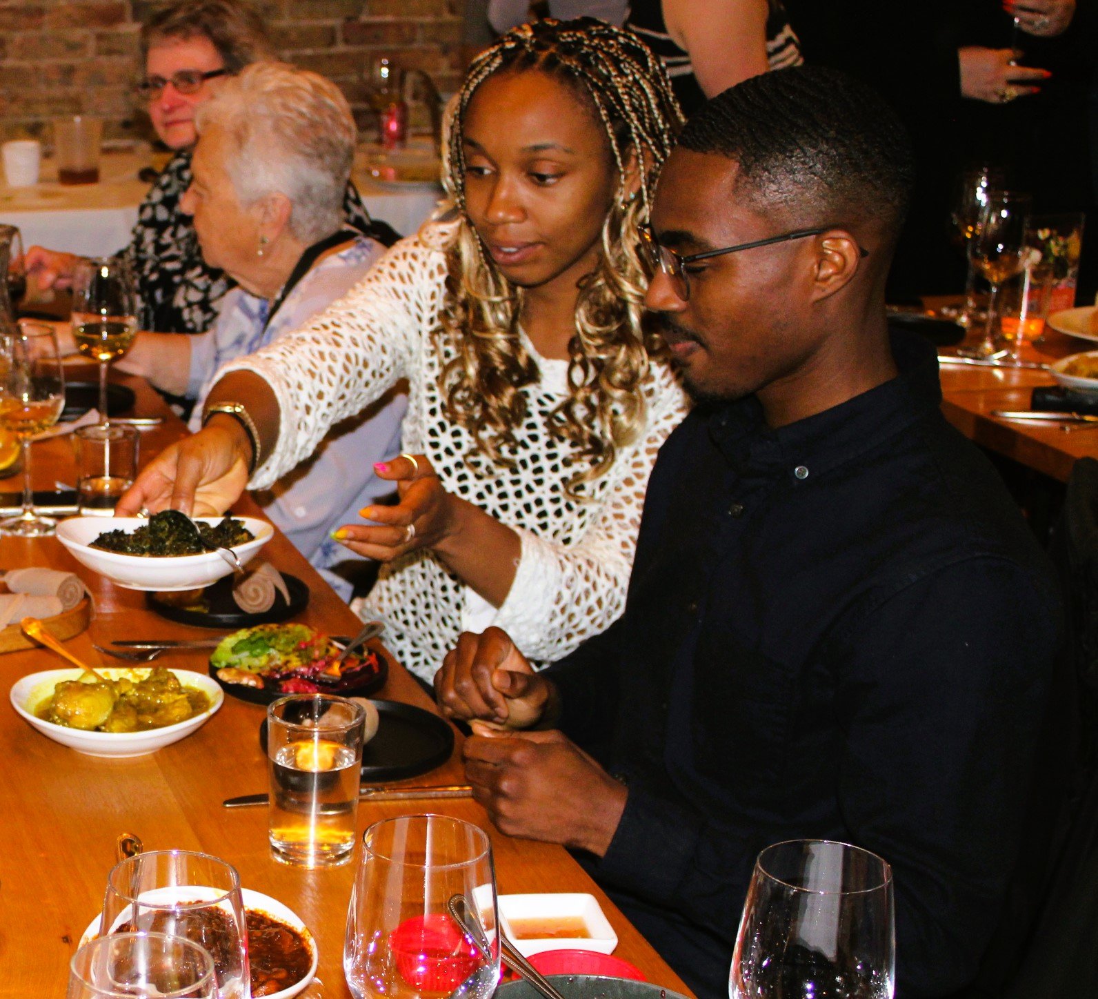 People dining at a restaurant, with a woman serving greens to a man, and others sitting at the table with plates and drinks.